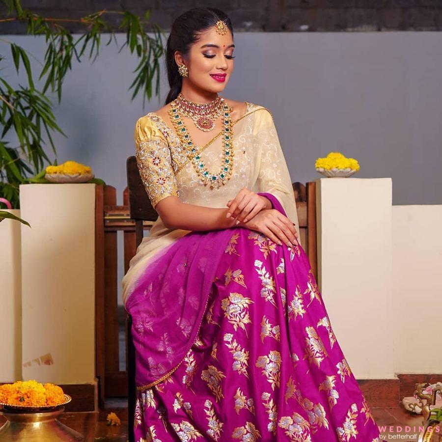 A beautiful woman wearing a purple sari and gold jewelry sits amidst lush potted plants.