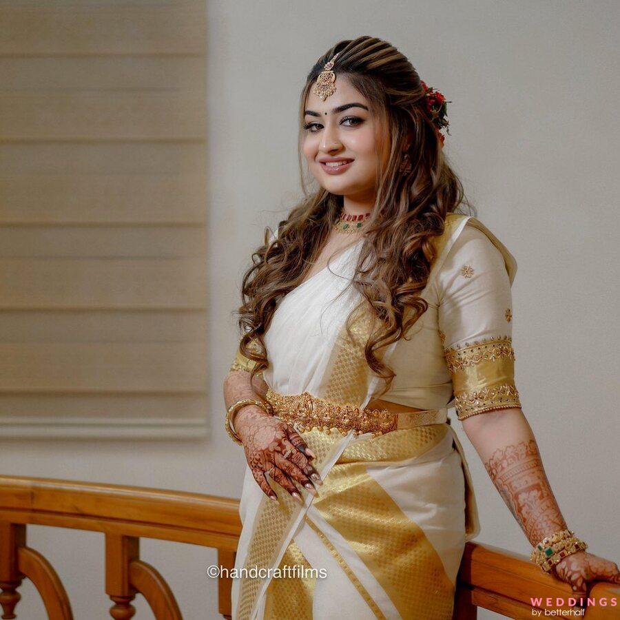 A beautiful woman in a gold-colored sari, standing on a wooden railing with a radiant smile.