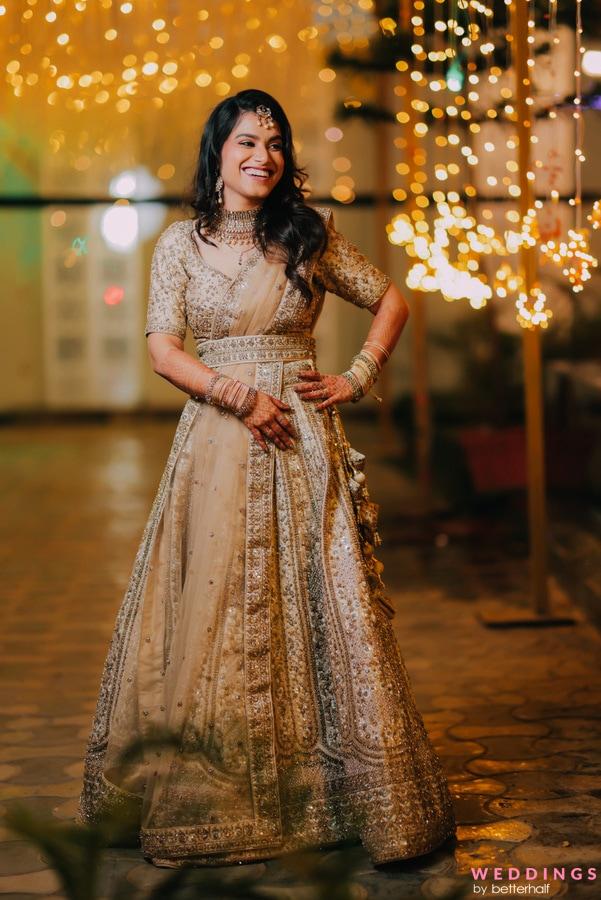 A stunning bride in a gold-colored lehenga stands amidst a walkway adorned with twinkling string lights.