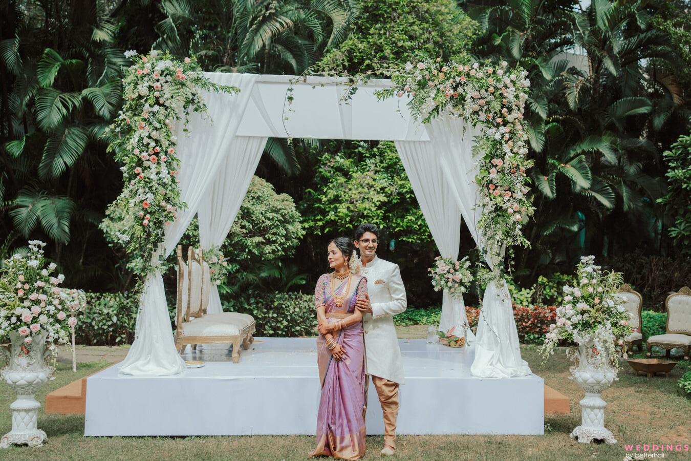 A couple posing under a white canopy in a lush green garden for their traditional Hindu wedding ceremony.